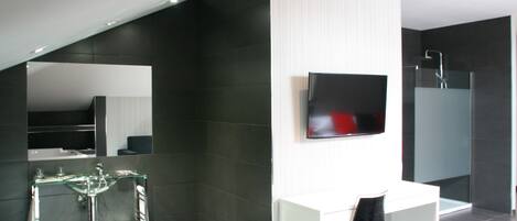 Un baño moderno con un lavabo de vidrio, un tocador blanco, un televisor de pantalla plana y una zona de ducha.