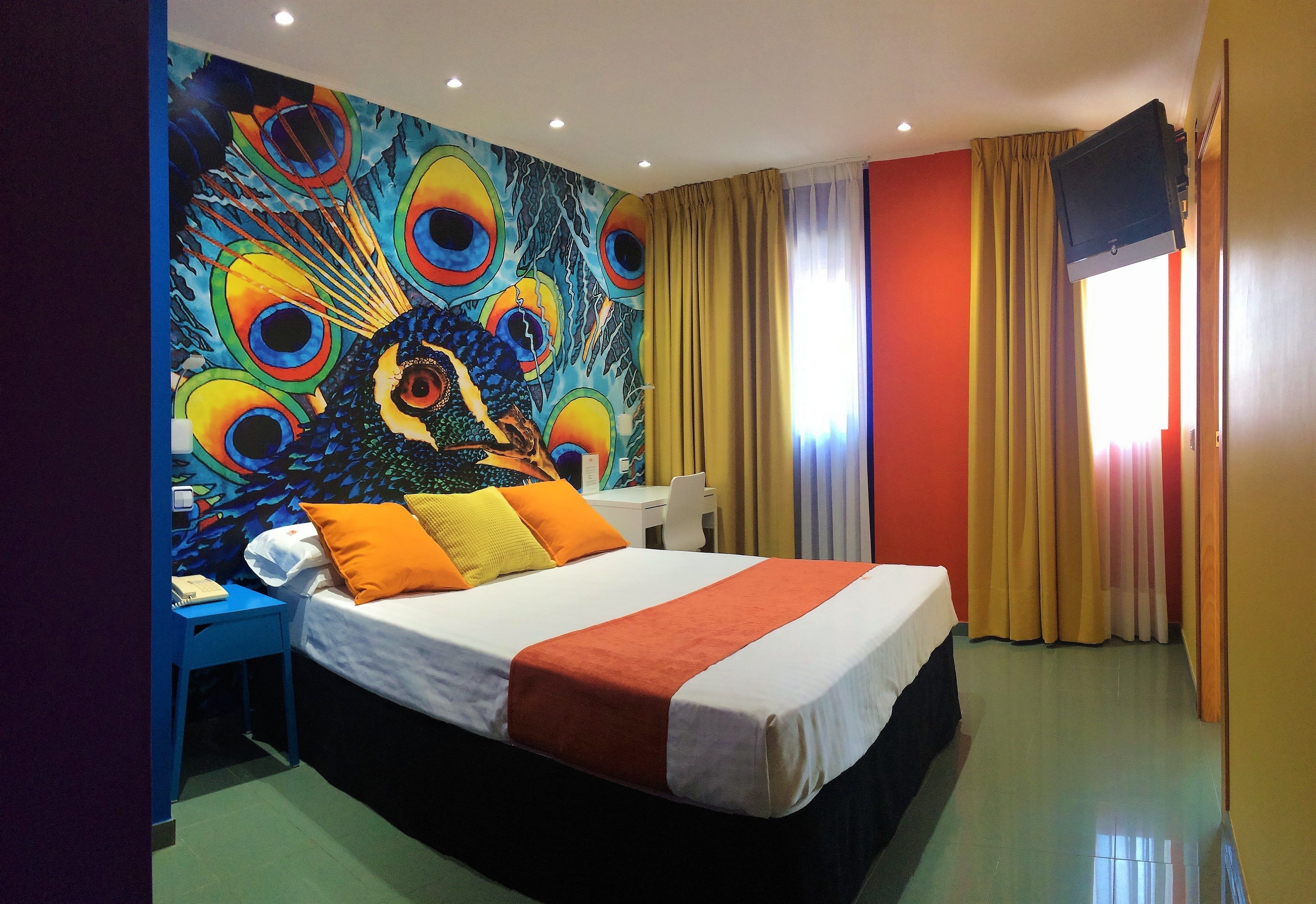 Habitación de hotel con un gran mural de un pavo real en la pared, una cama con ropa de cama naranja y blanca, un escritorio con una silla y un televisor montado en la pared.