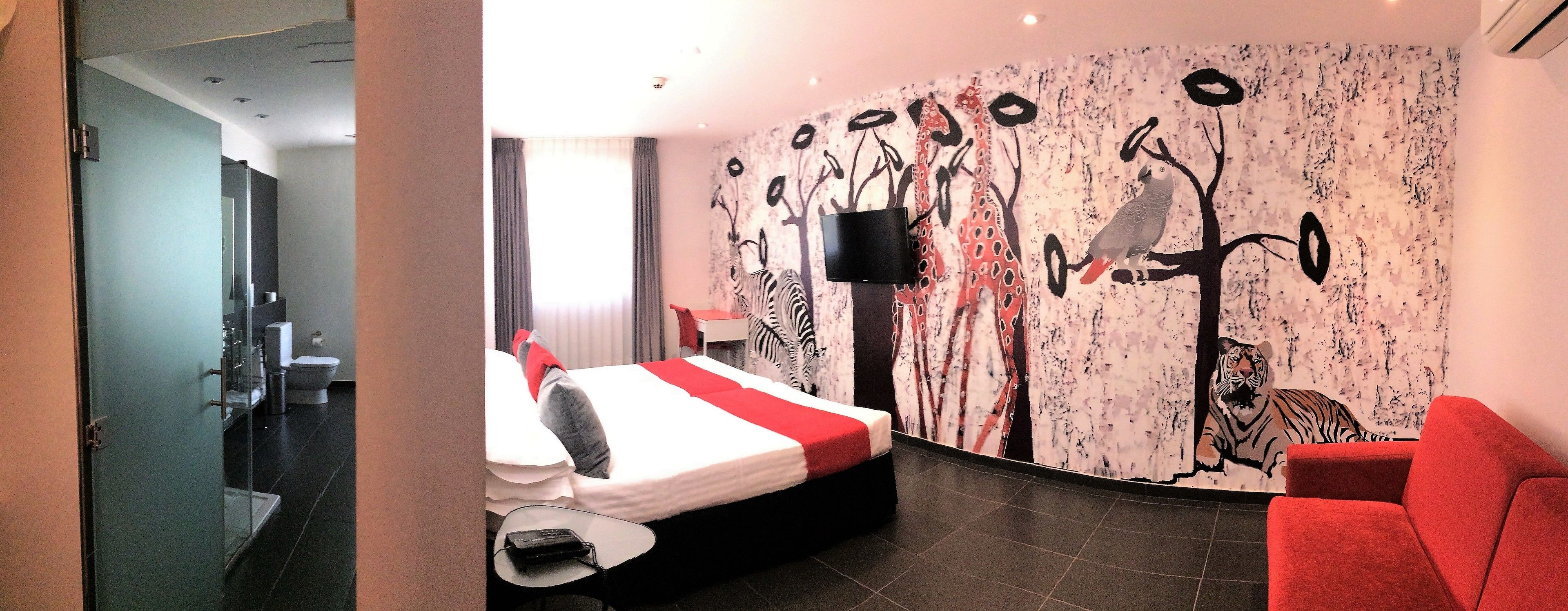 A hotel room with a bed, a red armchair, a wall-mounted TV, and a bathroom visible through an open door.