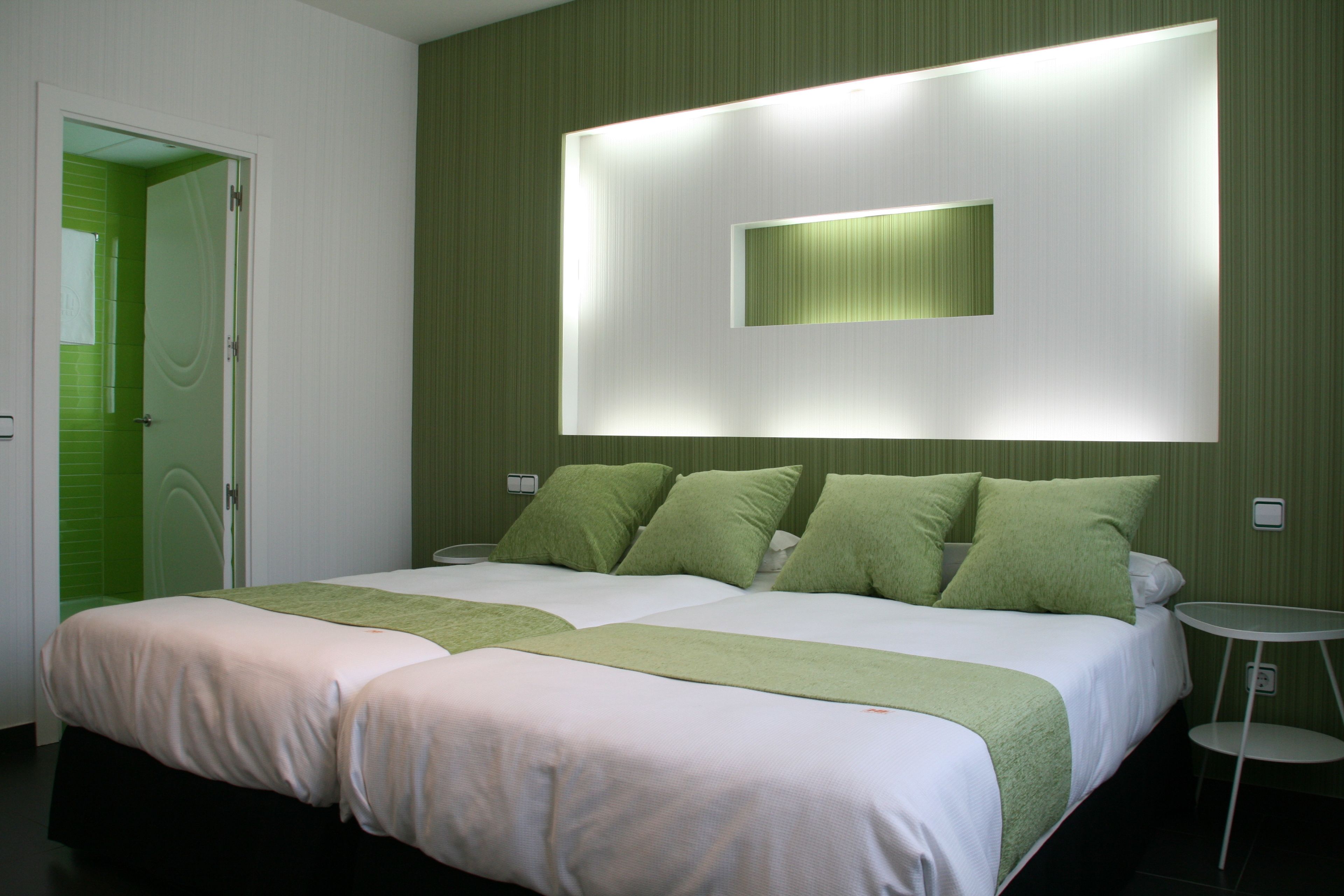A hotel room with two beds, a green accent wall, a bedside table, and a wall-mounted light fixture.