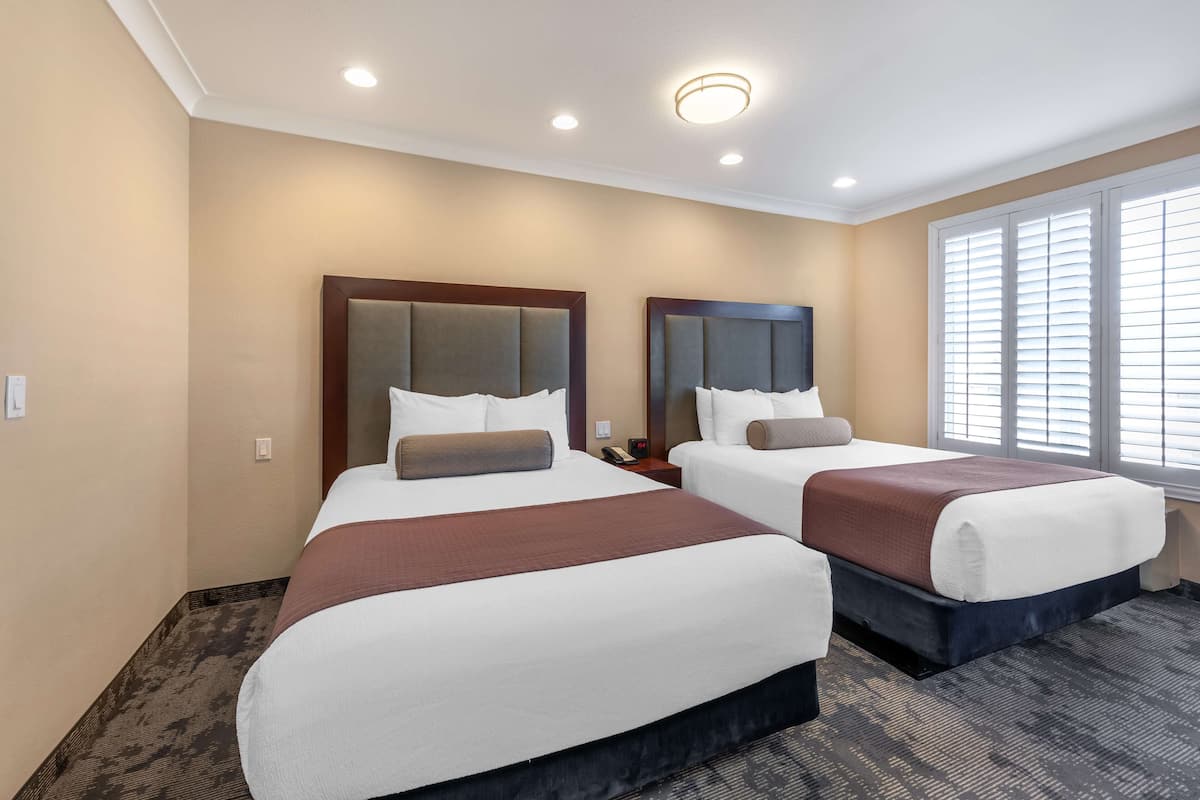 A hotel room with two beds, beige walls, a large window with blinds, and a carpeted floor.