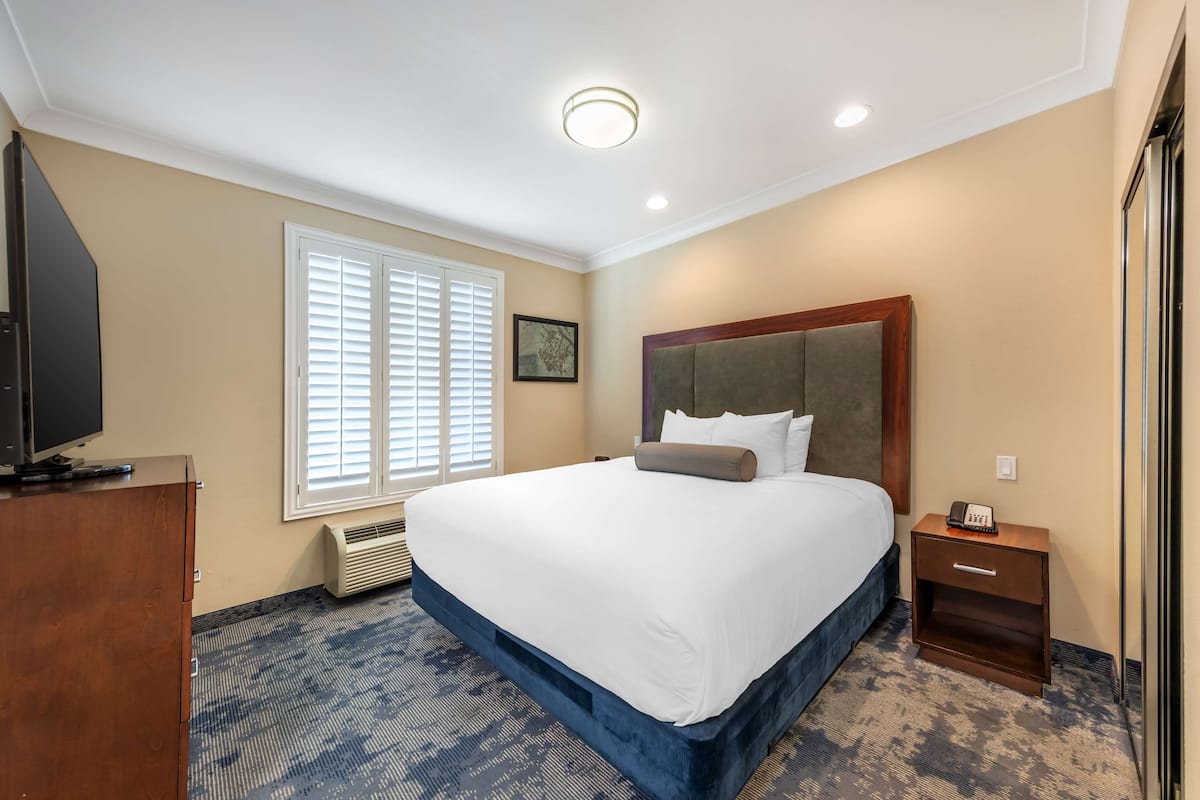 A hotel room with a large bed, a wooden nightstand, a television on a wooden dresser, and a window with blinds.