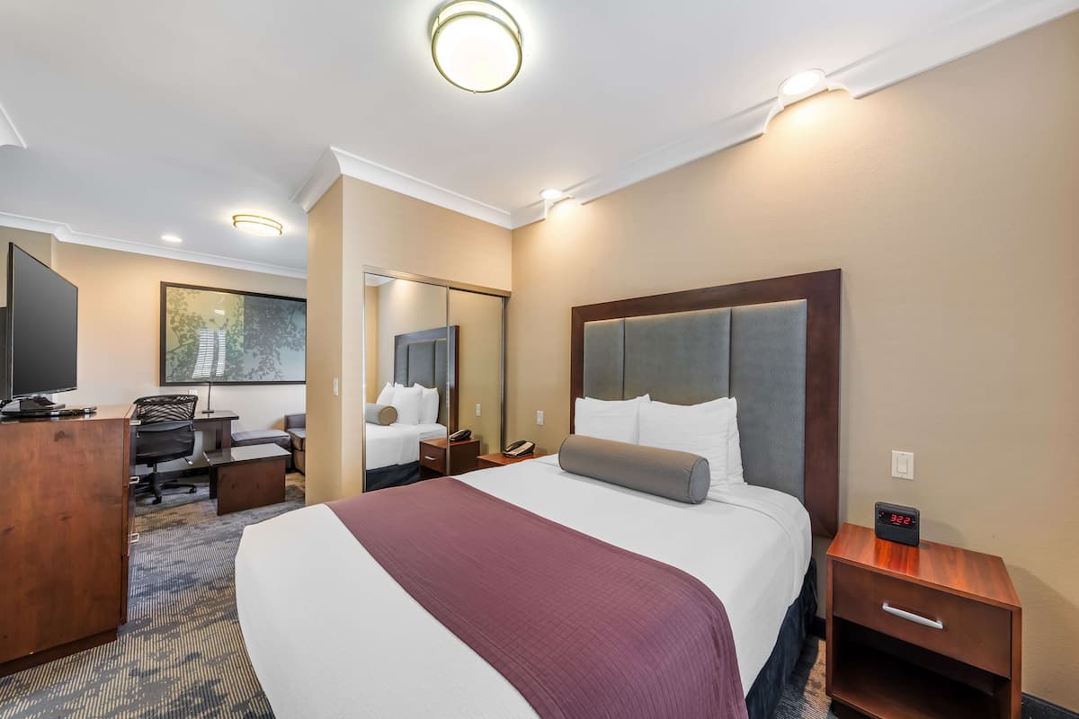 A hotel room with a large bed, a desk, a chair, a mirror, and a nightstand with a clock.