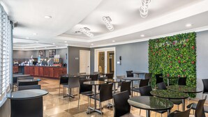 Un café moderno con mesas redondas y sillas negras, una pared verde con flores y una barra con máquinas de café.