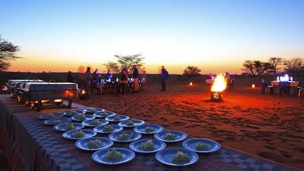 Ein Wüstencamp mit Lagerfeuer, langem Tisch mit Tellern und einem Wohnwagen bei Sonnenuntergang.