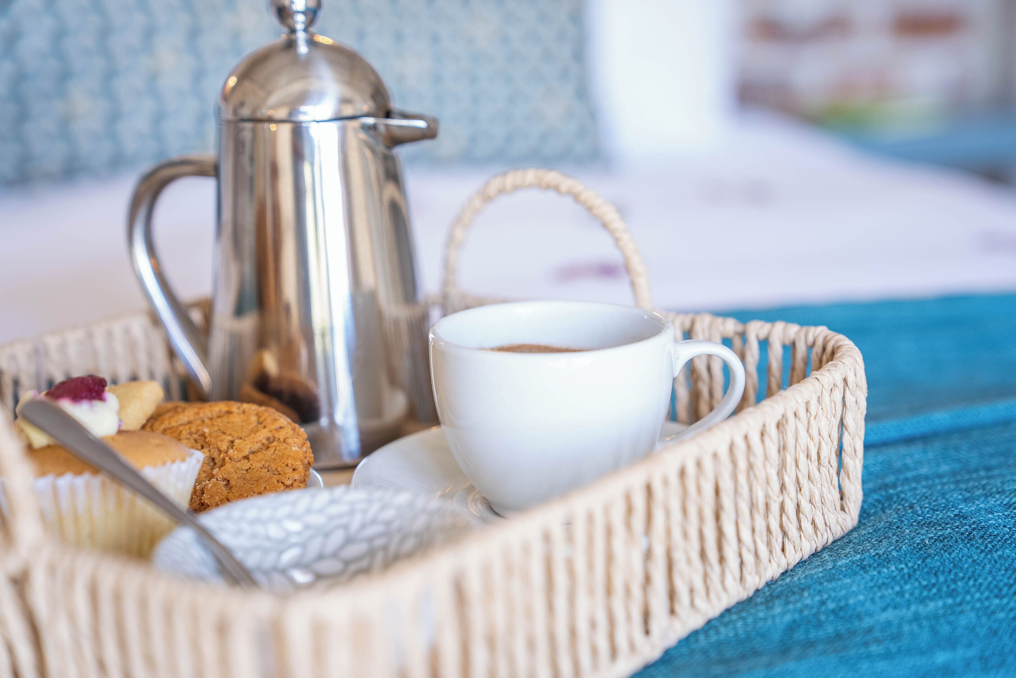 A wicker basket containing a white cup, a silver coffee pot, a cookie, and a pastry.