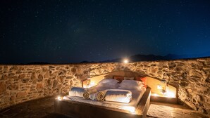 Una habitación de paredes de piedra con una cama, vista al cielo nocturno y un farol.