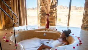 A person relaxing in a large bathtub filled with bubbles and rose petals, with a stone wall and a view of a desert landscape. - Le Mirage Resort and Spa (Sesriem)
