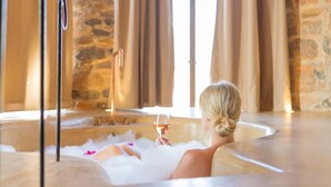 Una persona relajándose en una bañera, con una copa de vino, rodeada de paredes de piedra y cortinas.