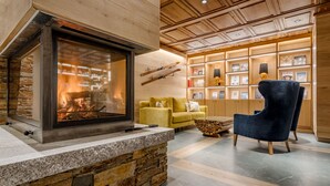 A cozy living area with a fireplace, a stone accent wall, a yellow sofa, a blue armchair, and a wooden bookshelf with framed pictures. - Hotel Chalet Bassibe (Naut Aran)