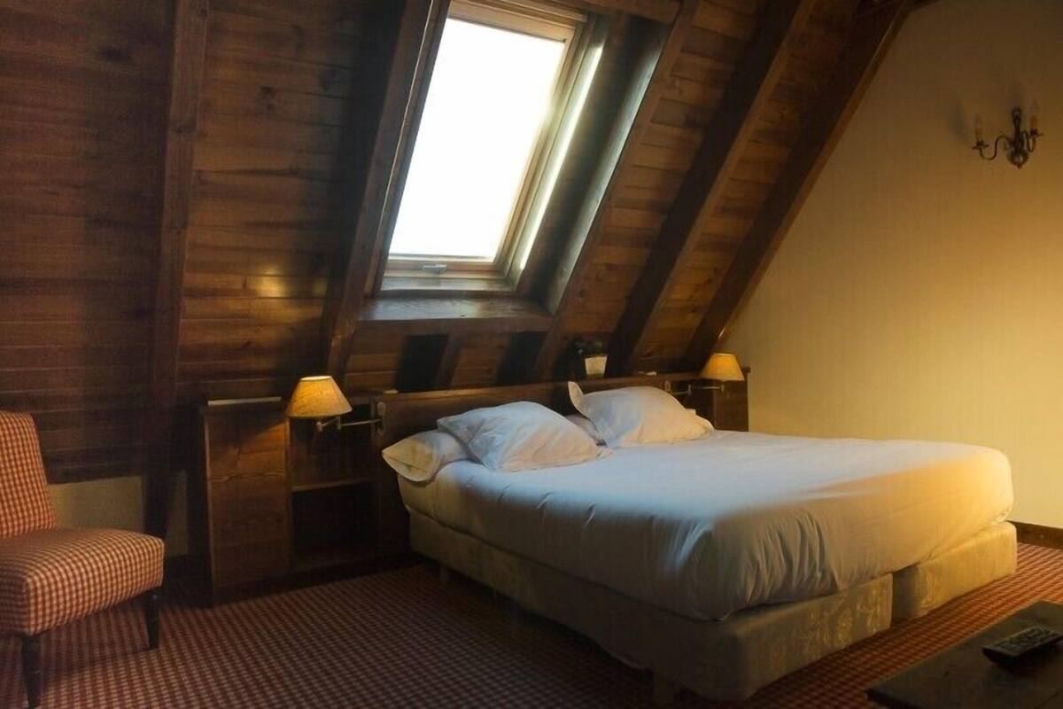 Ett sovrum med ett sluttande tak, en säng med vita sängkläder, två sänglampor, en rutig fåtölj och ett litet bord med en fjärrkontroll.