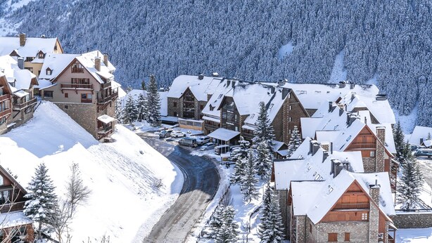 A snow-covered village with traditional wooden buildings, a winding road, and a backdrop of snow-laden trees and mountains. - Hotel Chalet Bassibe (Naut Aran)