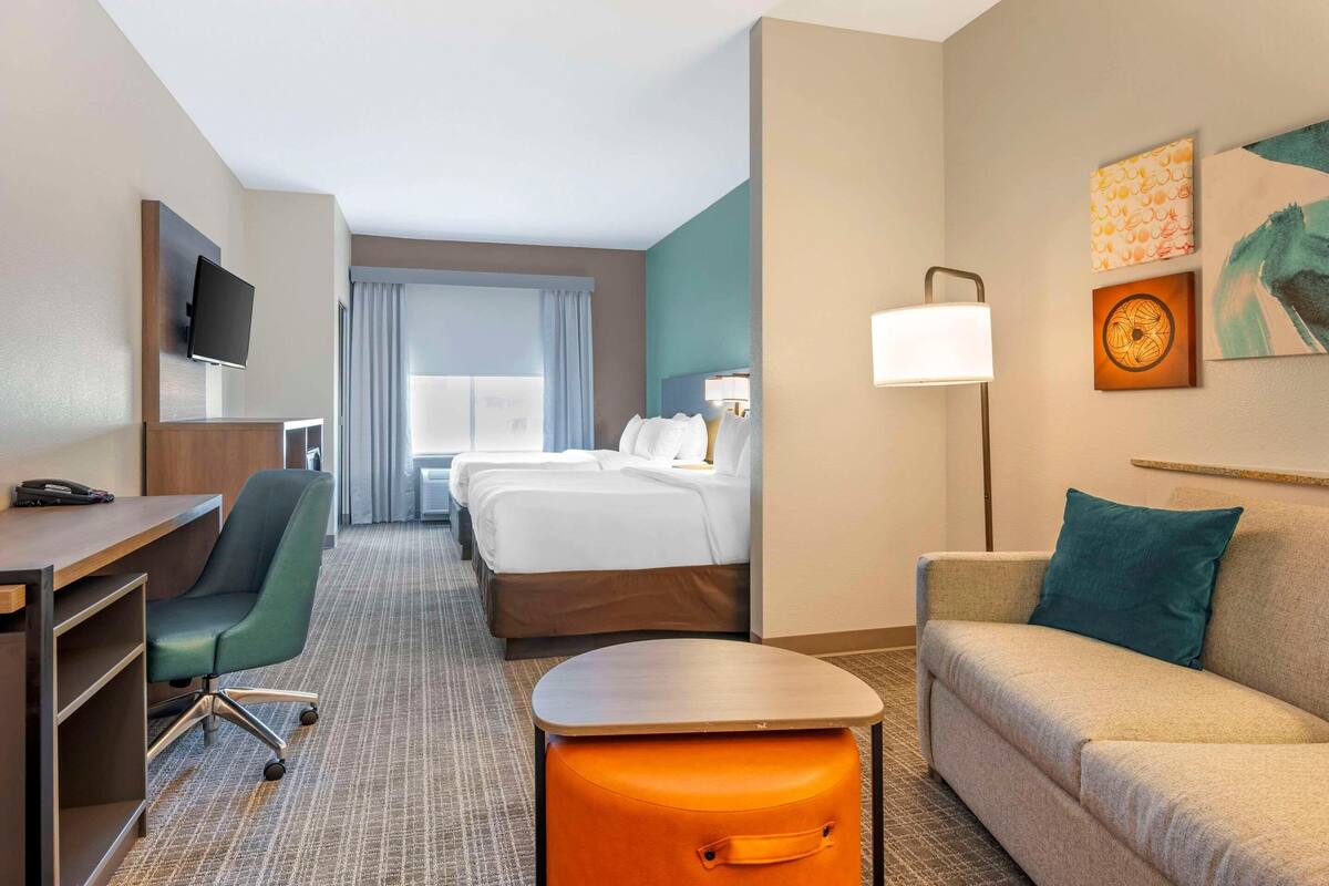 A hotel room with a desk, chair, bed, sofa, and a small orange ottoman.