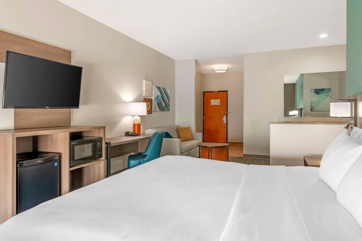 A hotel room with a bed, a desk with a computer, a microwave, a refrigerator, a TV, and a door with a sign.
