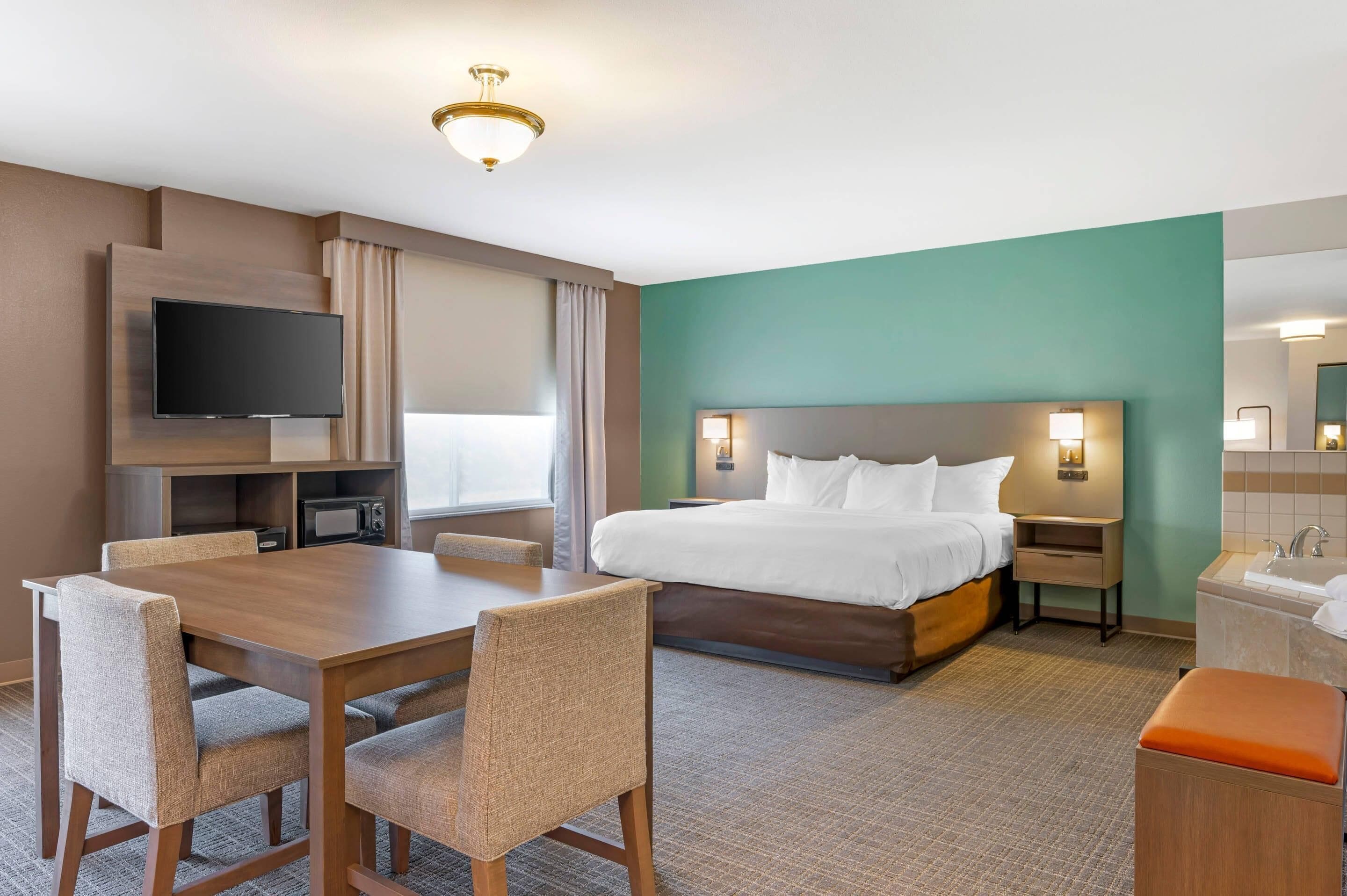 A hotel room with a bed, a TV, a dining table with chairs, a small kitchenette, and a bathroom.