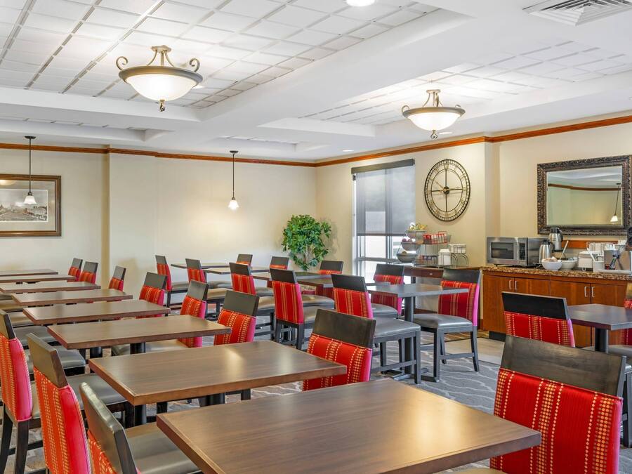 A hotel dining area with multiple tables and chairs, a clock, a plant, and a kitchen area with appliances.