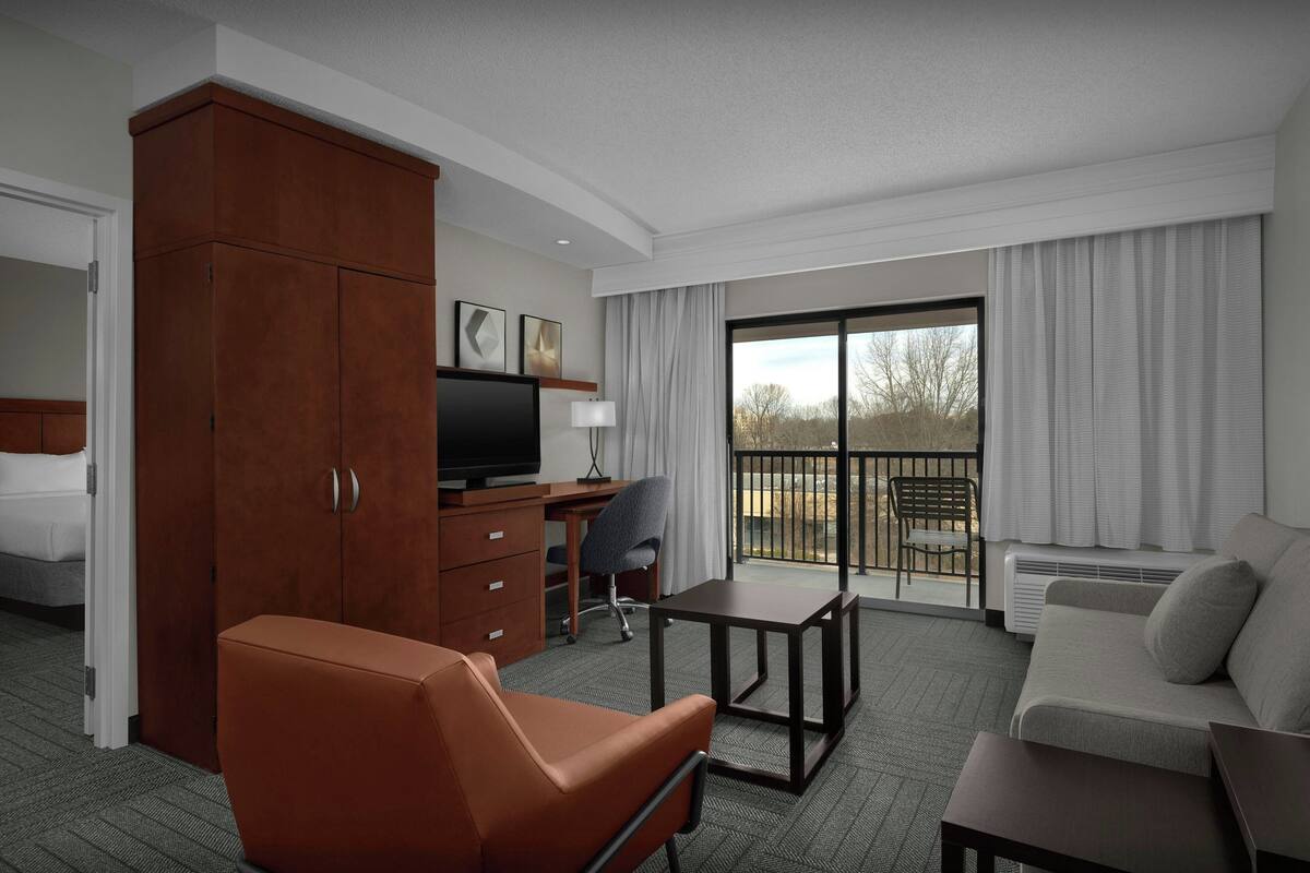 Ein Hotelzimmer mit braunem Ledersessel, einer Couch, einem Schreibtisch mit Computer, einem Fernseher und einem großen Fenster mit Vorhängen.