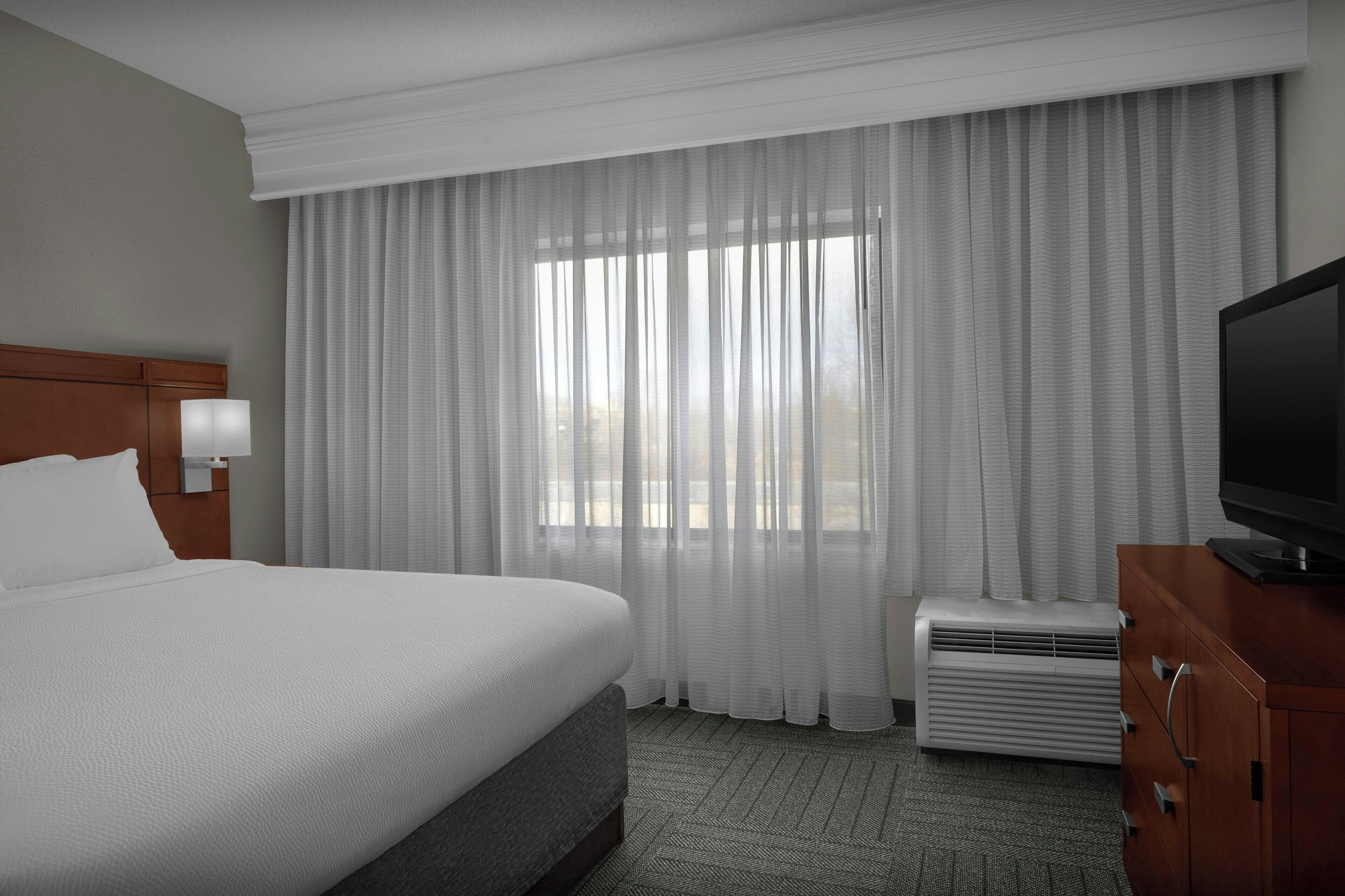 Habitación de hotel con una cama grande, televisión, calefacción y ventana con cortinas traslúcidas.