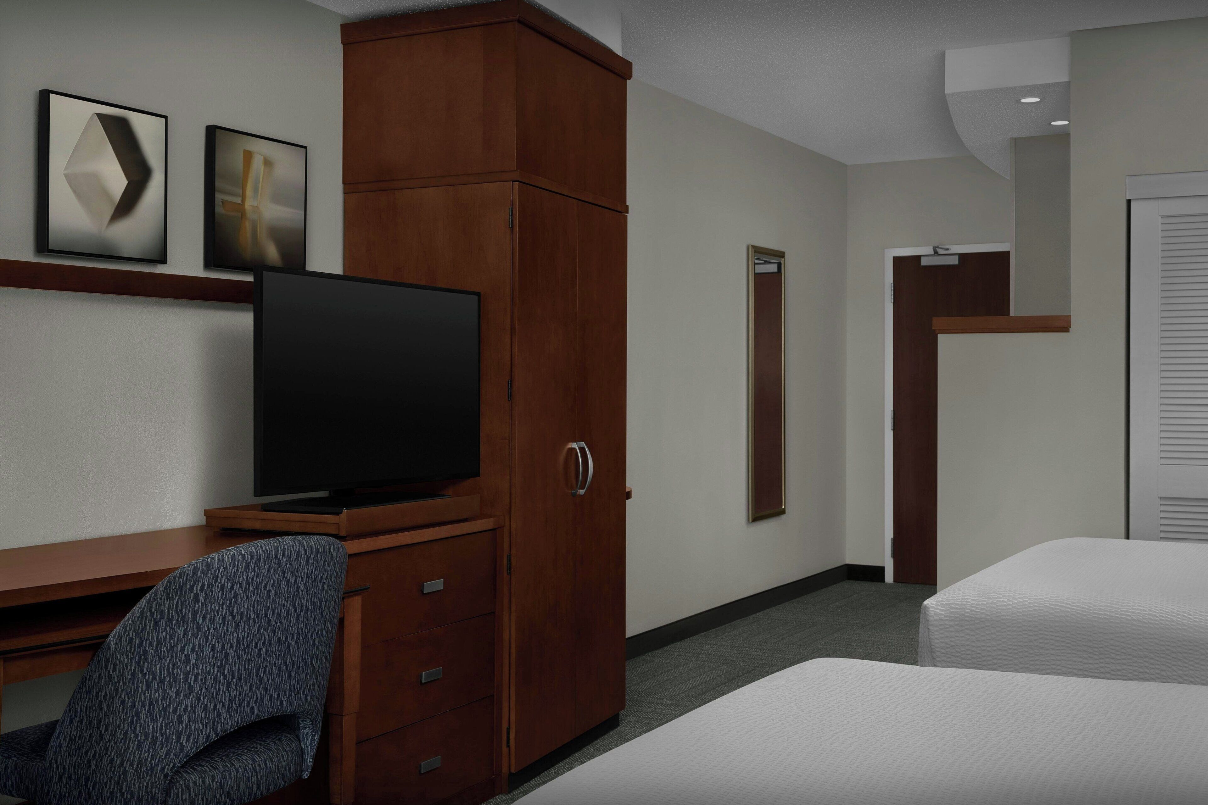 A hotel room with a wooden desk, a television, two beds, and artwork on the wall.