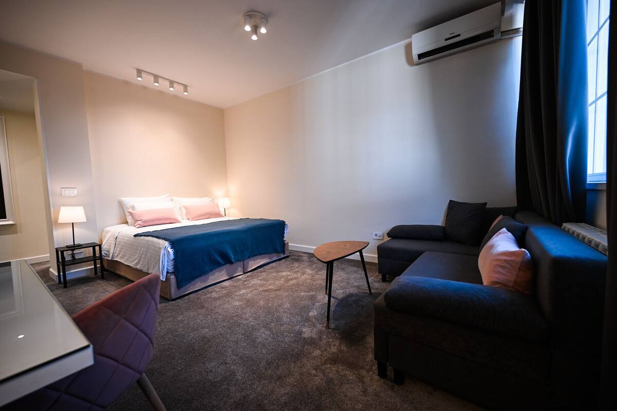 Quarto de hotel com uma cama, um sofá, uma mesinha, uma escrivaninha e um aparelho de ar condicionado.