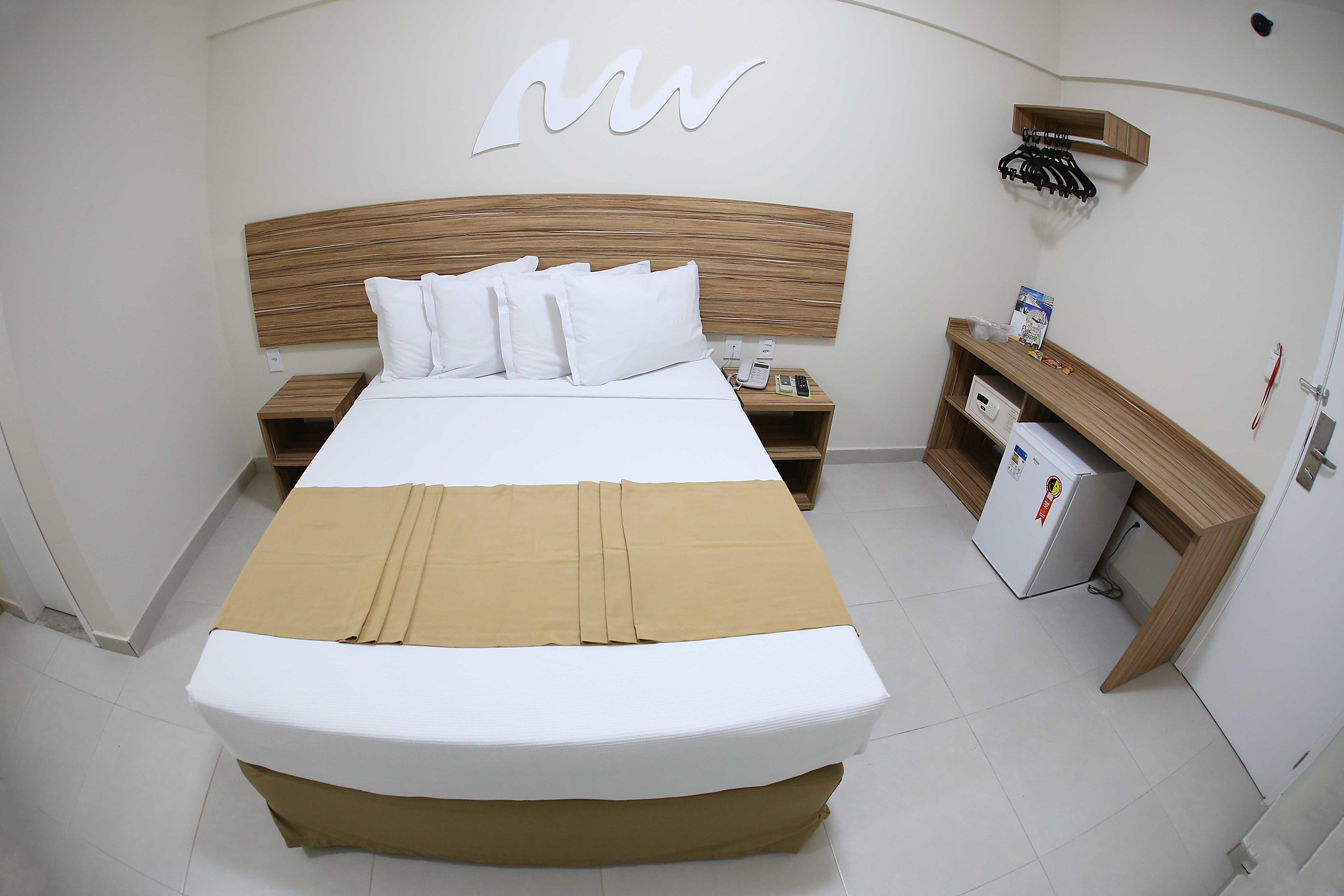 Una cama bien hecha con ropa blanca y falda marrón, un cabecero de madera, una mesita de noche con un teléfono y una cajita, y un perchero en la pared.