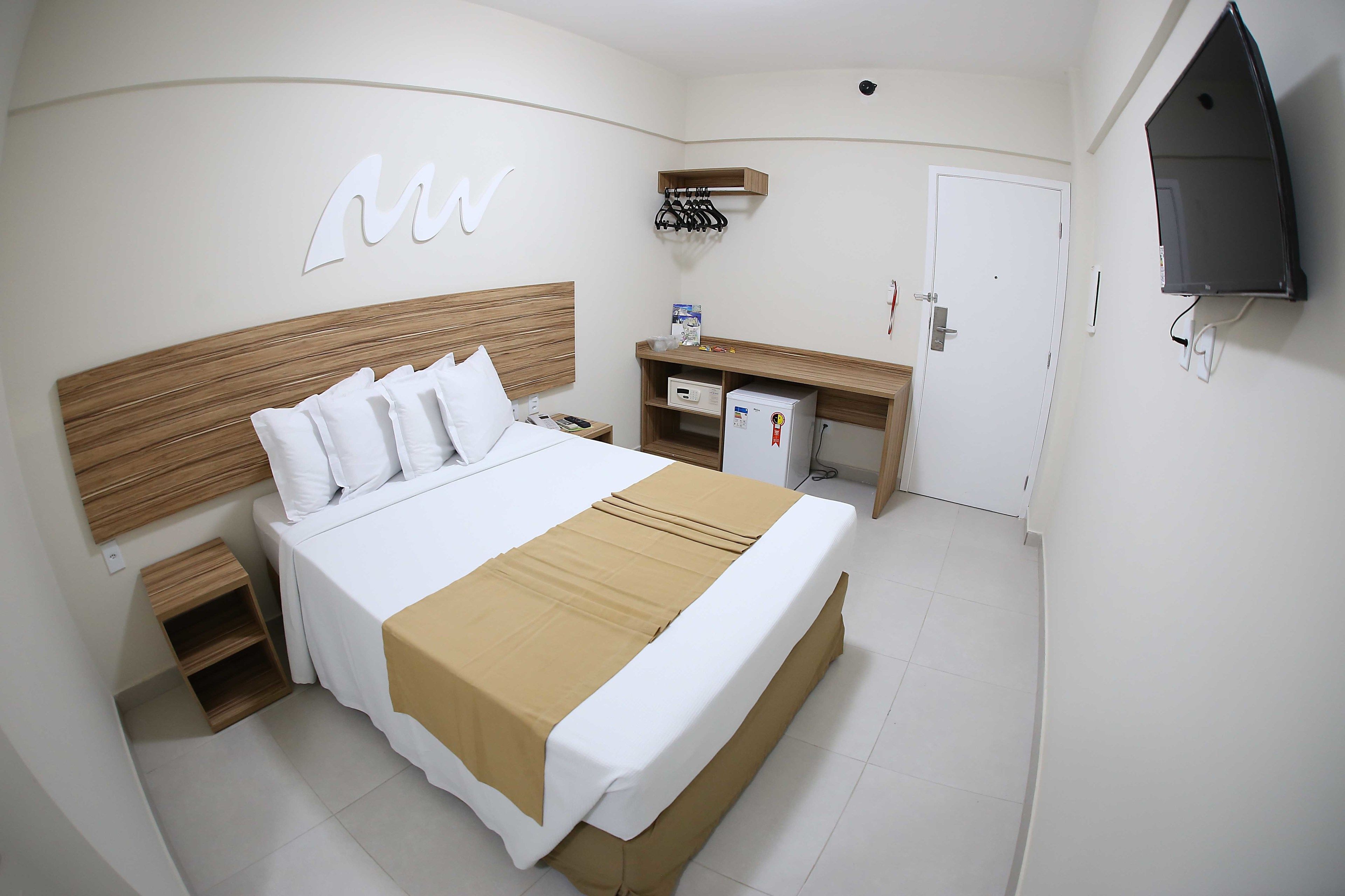 A hotel room with a bed, bedside table, a small shelf, a desk with a chair, a TV mounted on the wall, and a door.