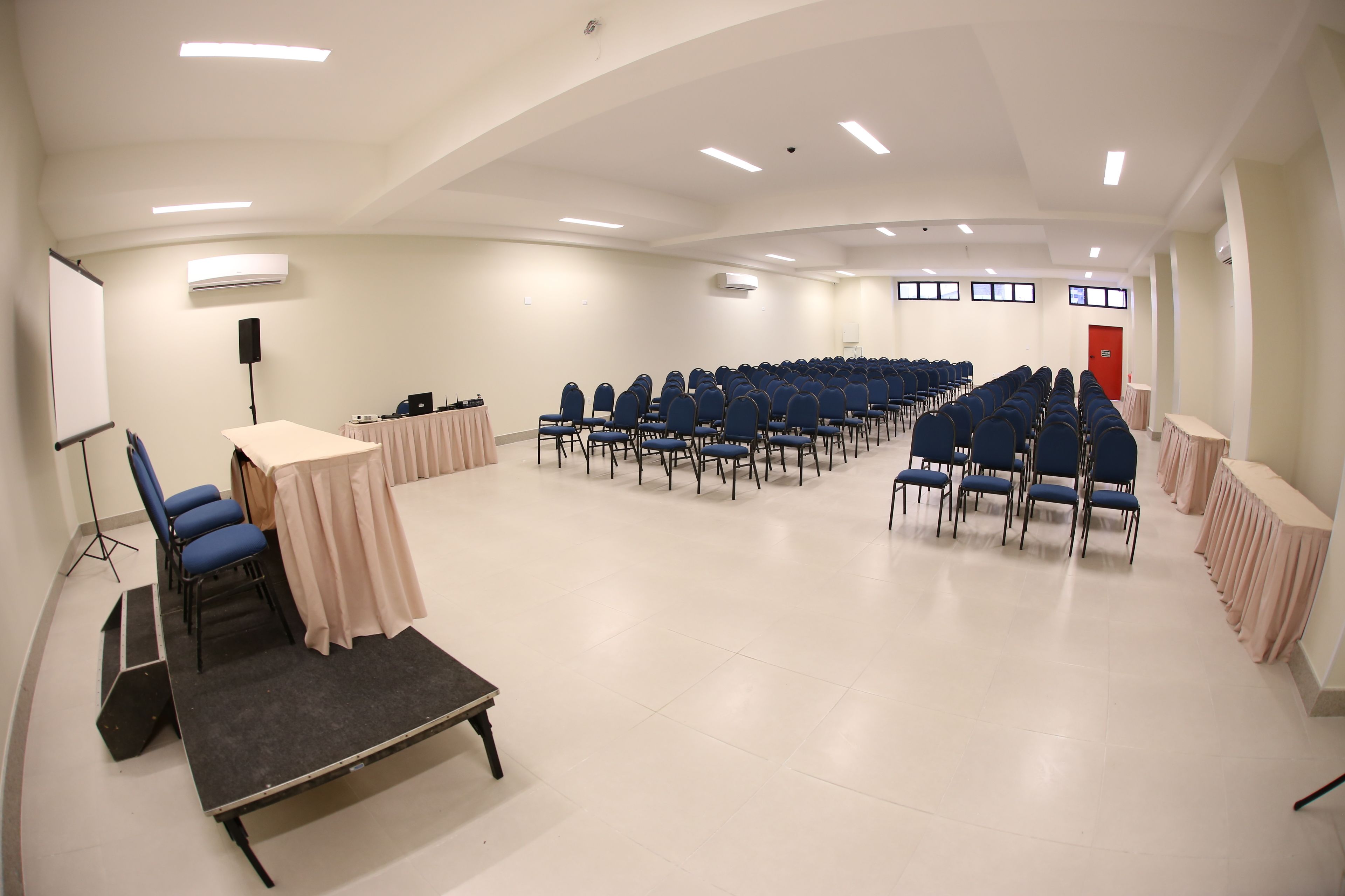 Una sala de conferències amb files de cadires blaves, un escenari amb un podi i una pissarra blanca.