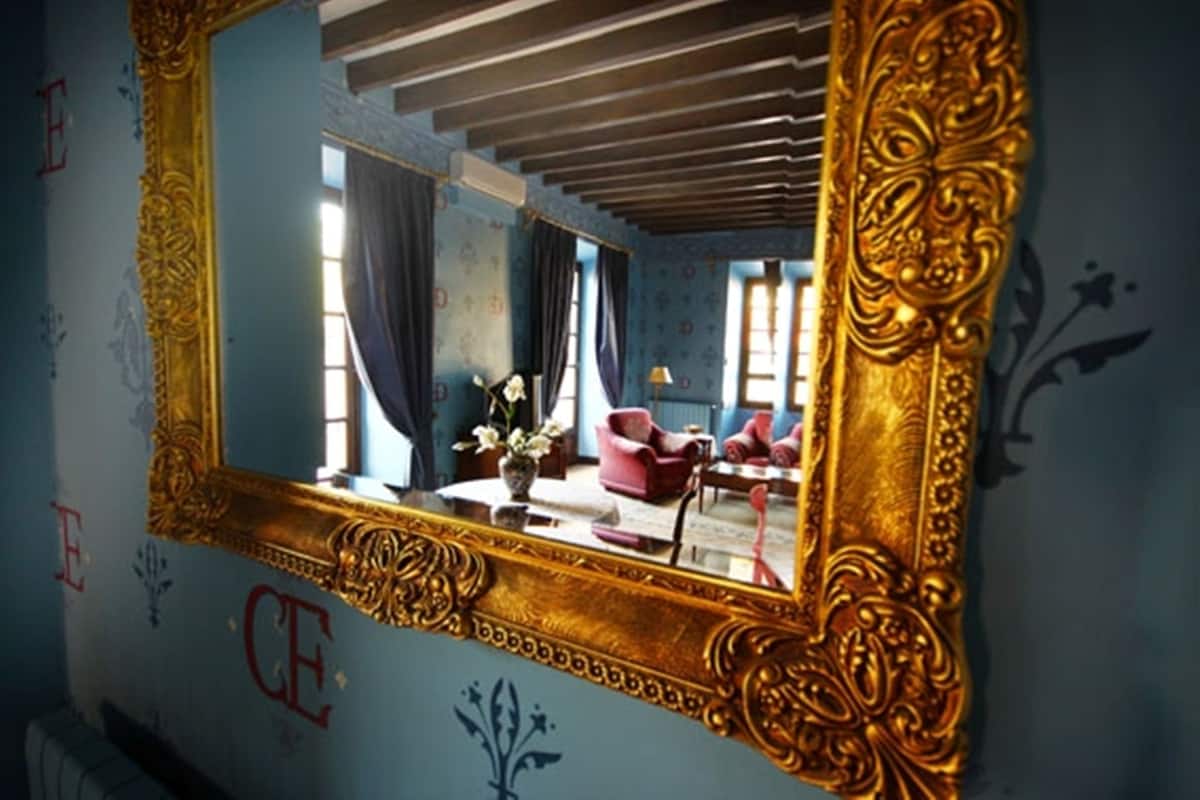Ein Zimmer mit einem großen, verzierten Spiegel, einer blauen Wand mit floralem Muster und einer Decke mit freiliegenden Balken.