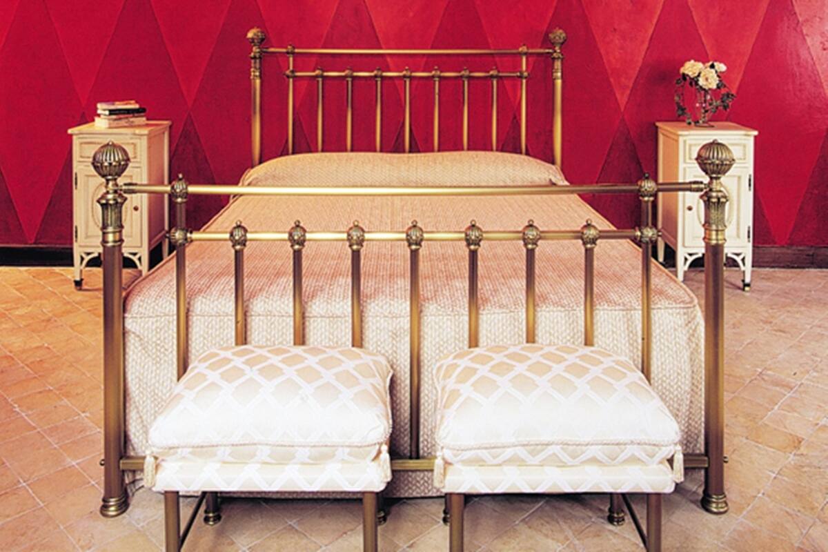 Ein Bett mit Metallgestell und zwei passende Fußstützen, an Nachttischen mit Lampen angelehnt, vor einer roten Wand mit Rautenmuster.