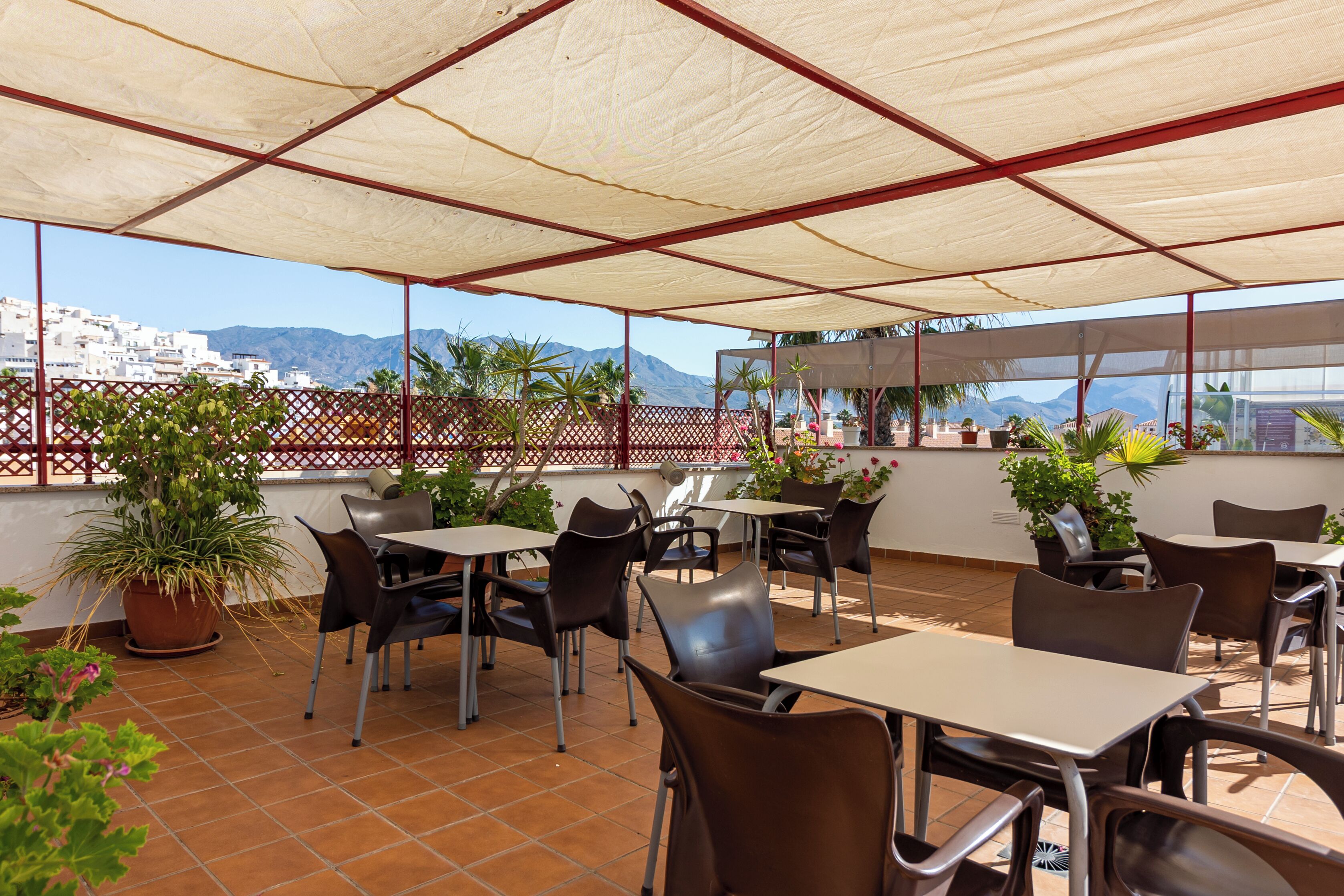 Un patio al aire libre con mesas y sillas, plantas en macetas y vistas a un pueblo costero y montañas.