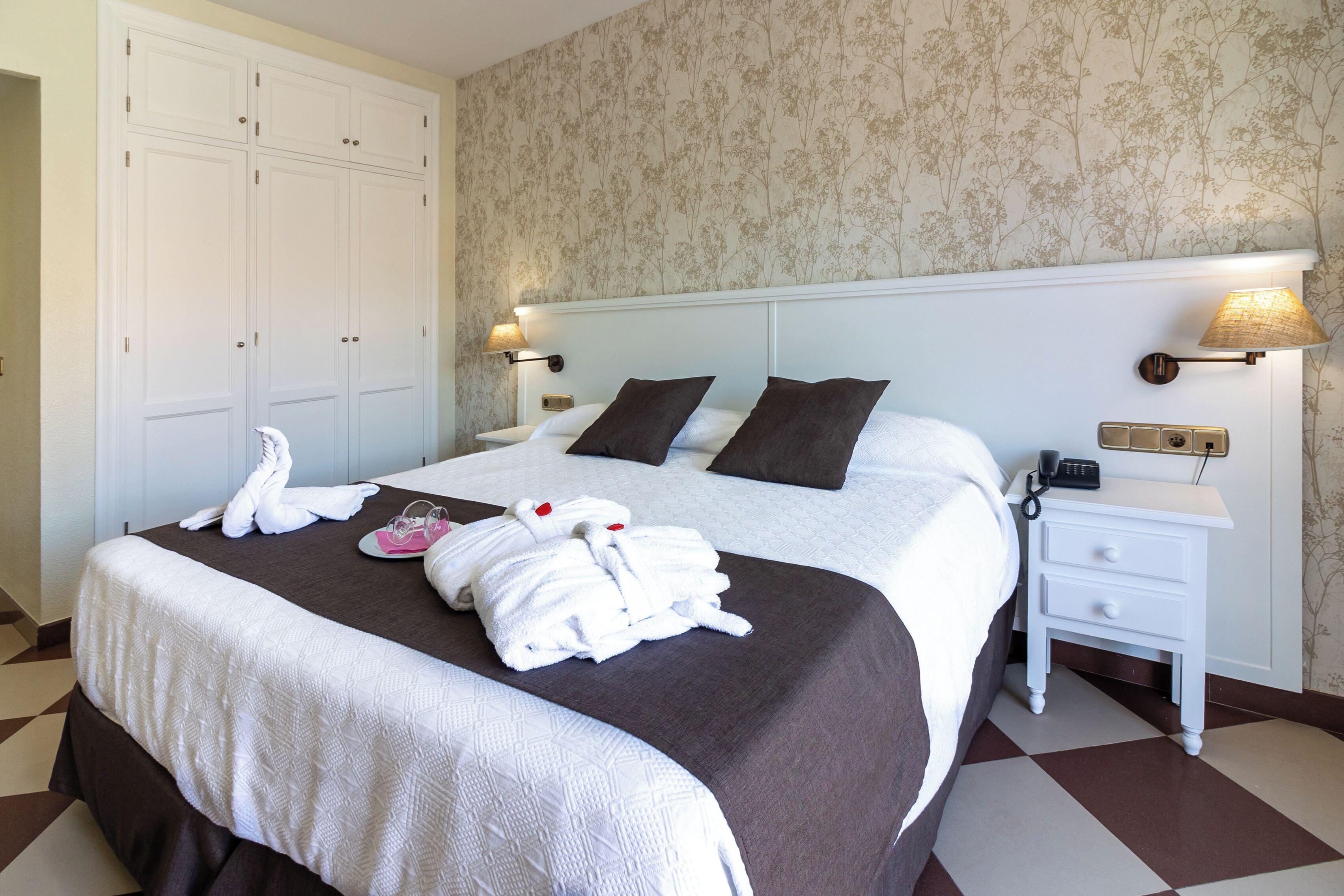 A neatly made bed with a brown headboard, white bedding, and two pillows. There are two bedside lamps, a white nightstand, and a wall-mounted telephone.