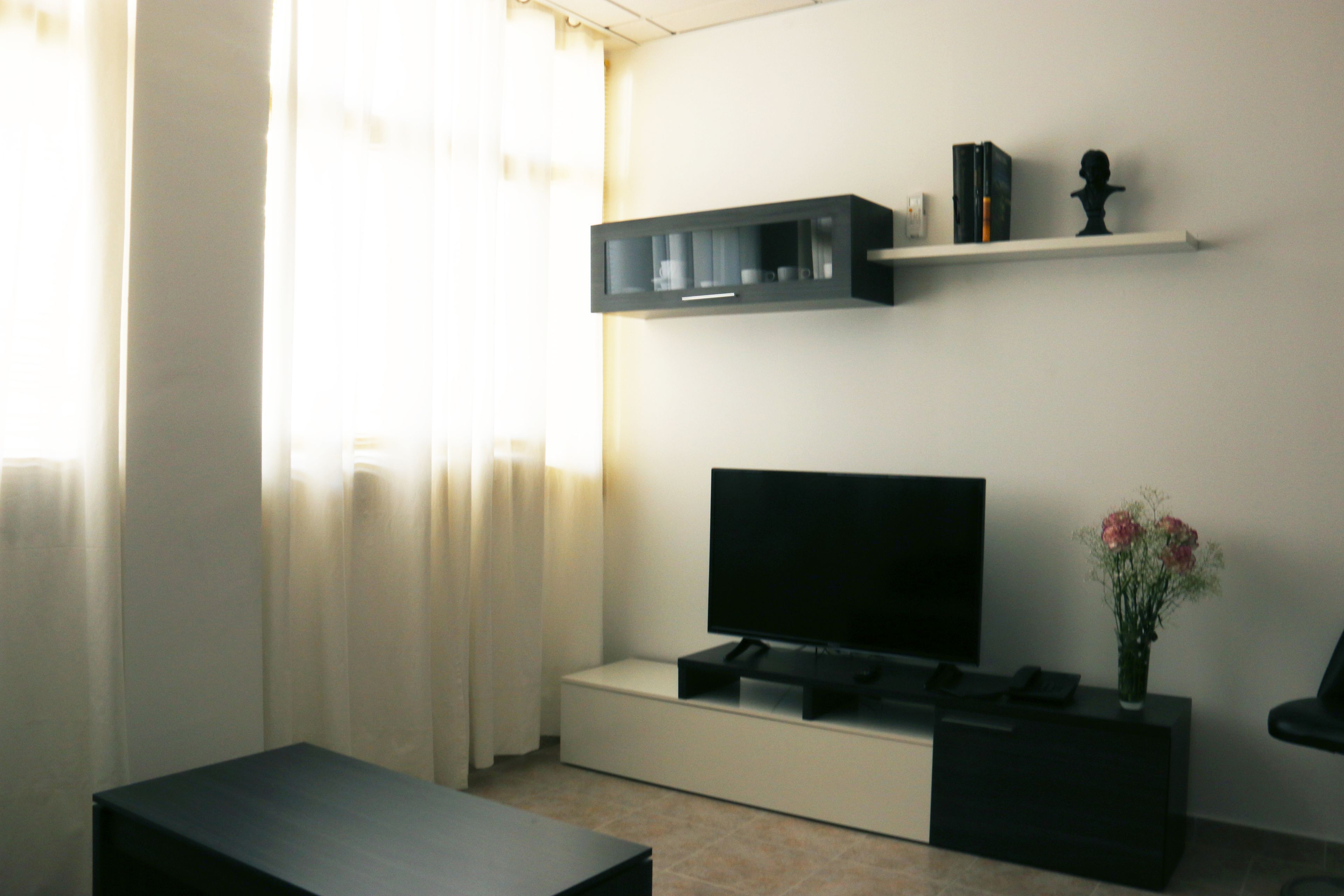 A modern living room with a flat-screen TV on a stand, a black cabinet above it, and a small table with a vase of flowers.
