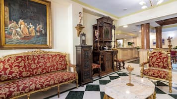Un amplio vestíbulo con suelo a cuadros, muebles ornamentados en rojo y dorado, un cuadro clásico de gran tamaño y un detallado aparador de madera.