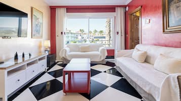 Una habitación de hotel con piso a cuadros, un sofá blanco, una mesita de noche roja, un televisor y un cuadro en la pared.