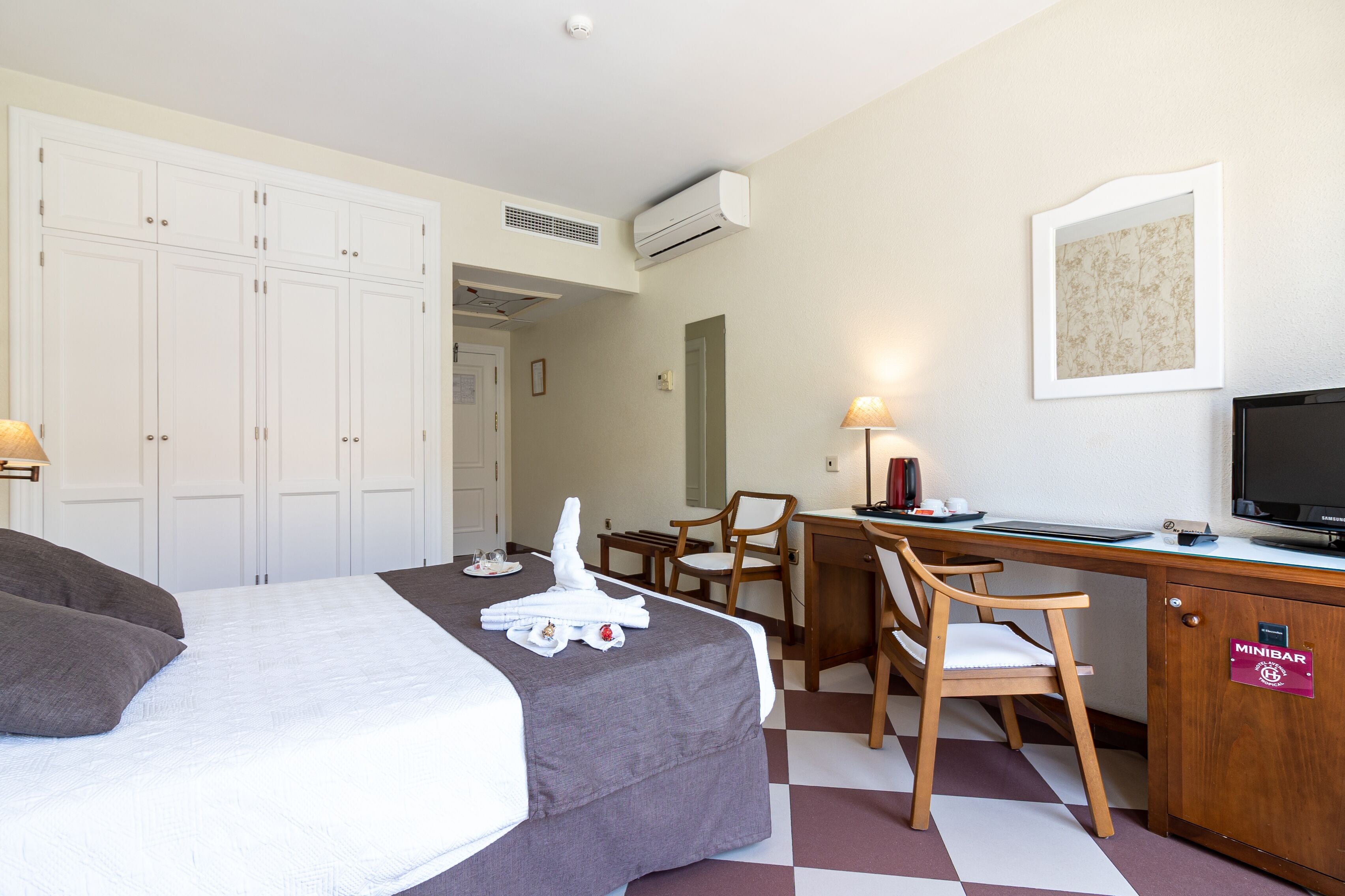 Quarto de hotel com cama, escrivaninha, cadeira, televisão e minibar.