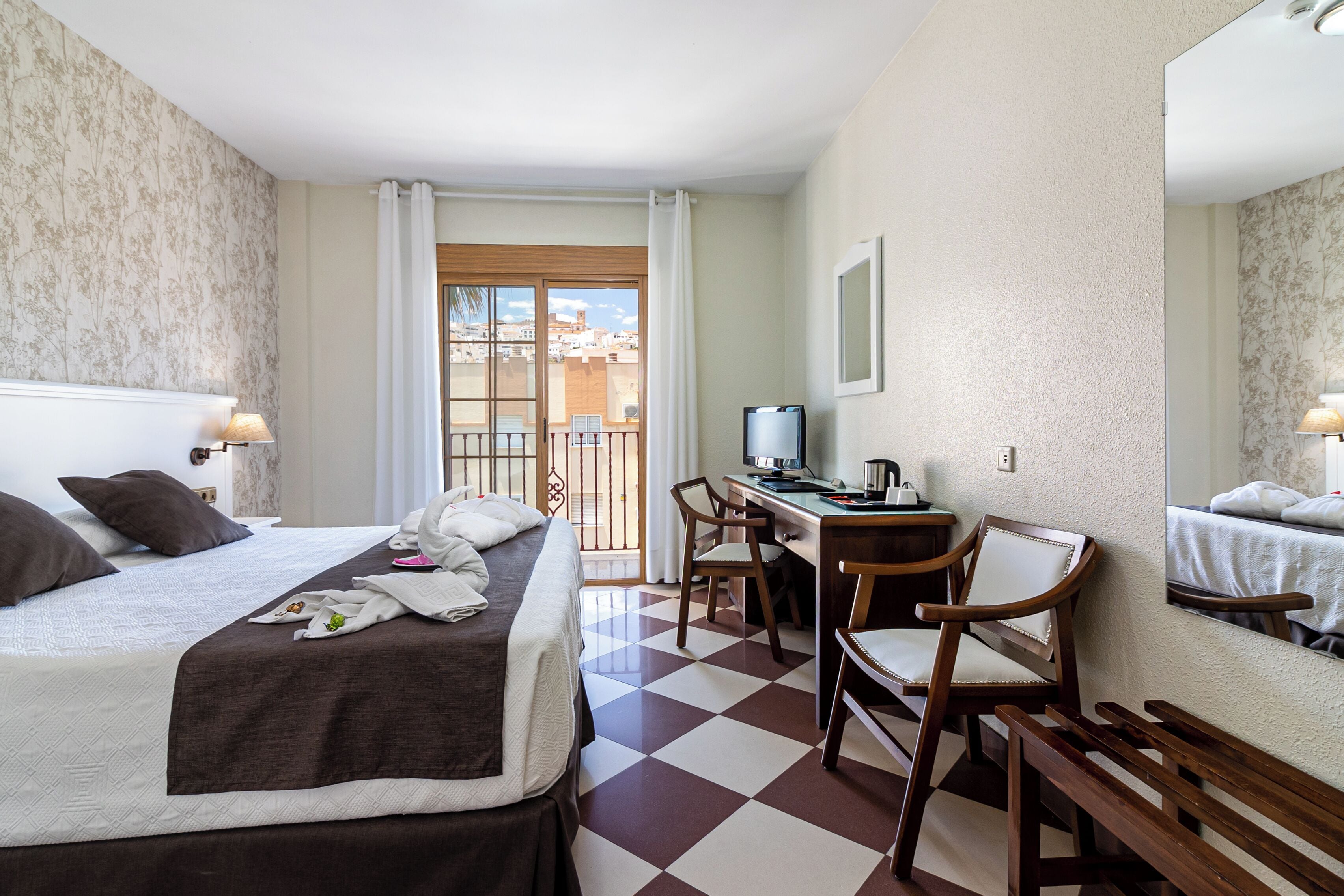 A hotel room with a bed, desk, chairs, and a balcony with a view.