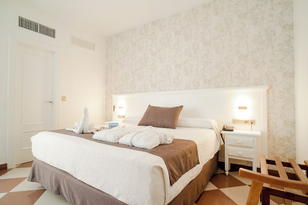 A neatly made bed with a brown headboard, a bedside table with a lamp, and a wooden chair.