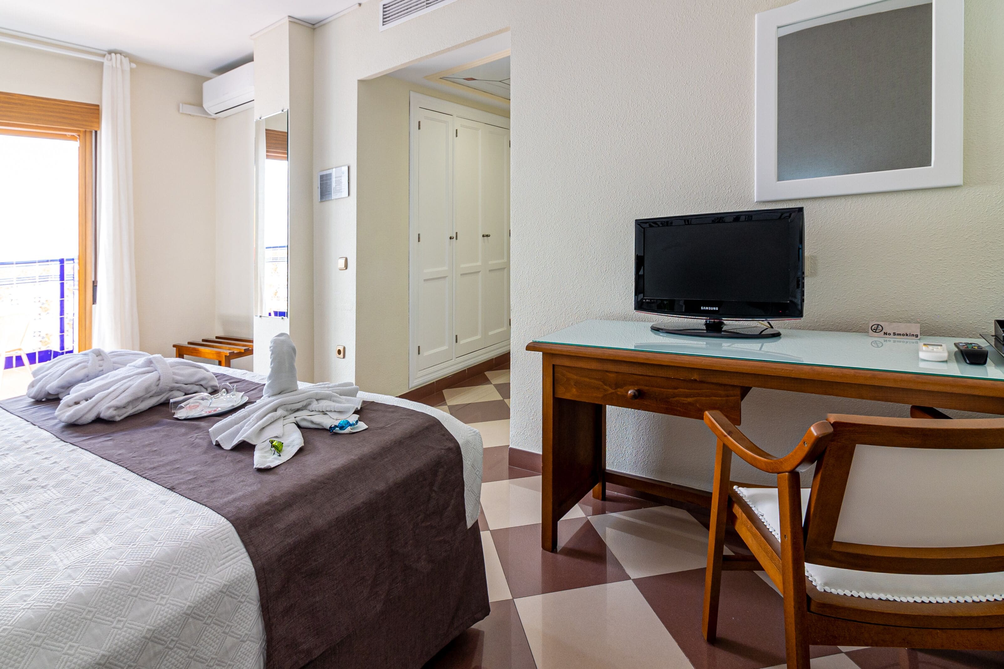 A hotel room with a bed, a desk with a computer, a chair, and a view of a balcony.
