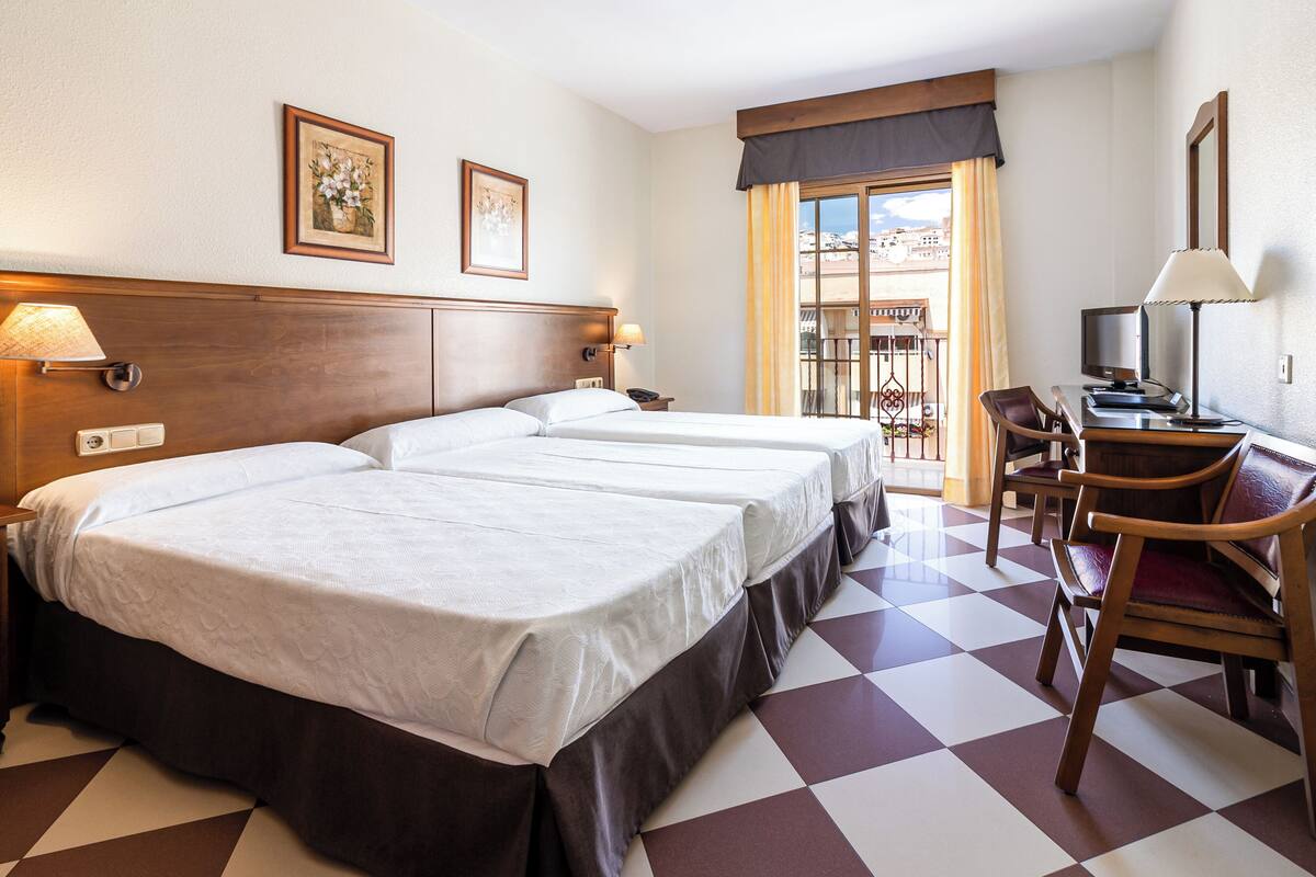 A hotel room with two beds, a desk with a chair, a television, and a balcony with a view.