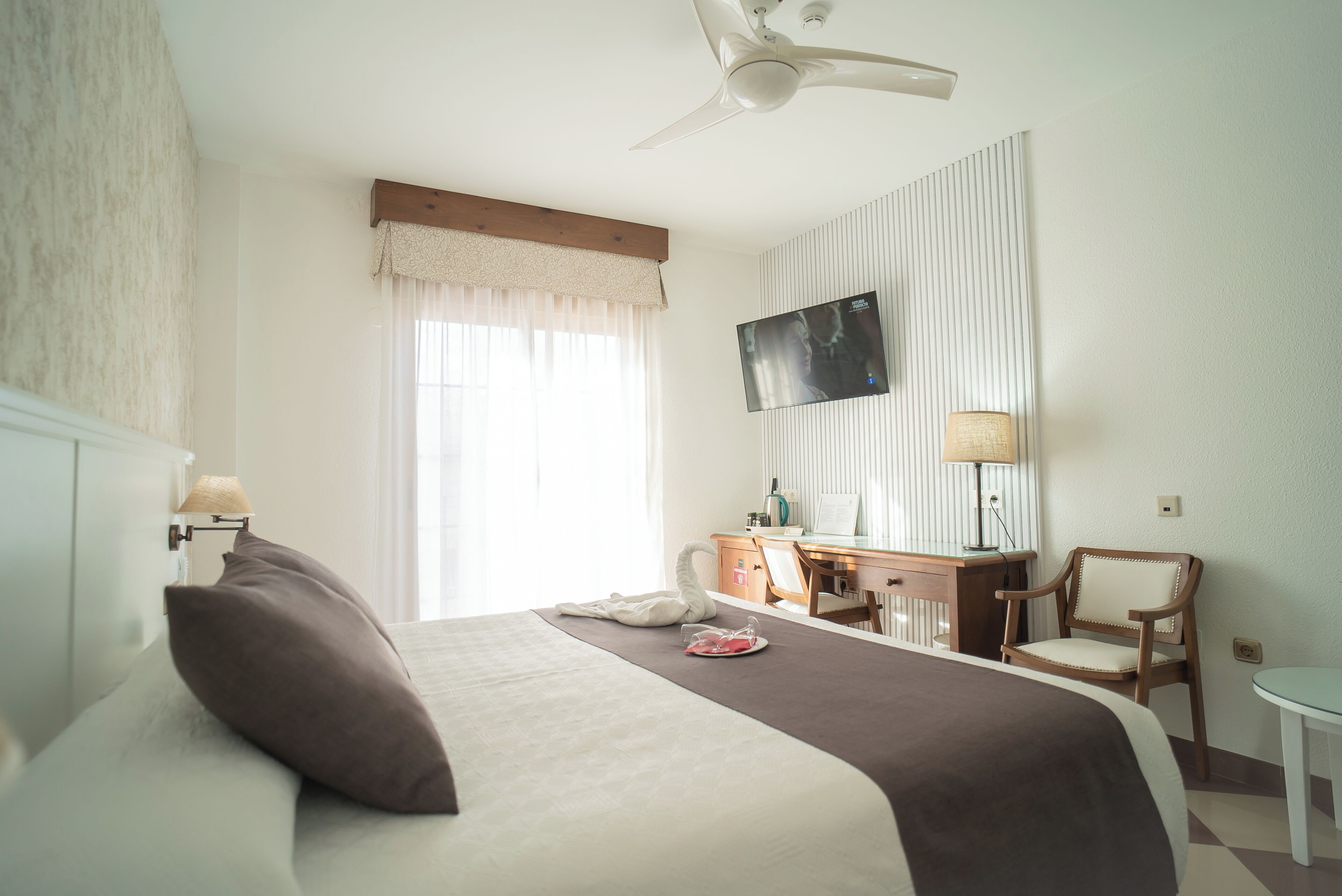 Una habitación de hotel con cama, escritorio, silla, televisor y ventilador de techo.