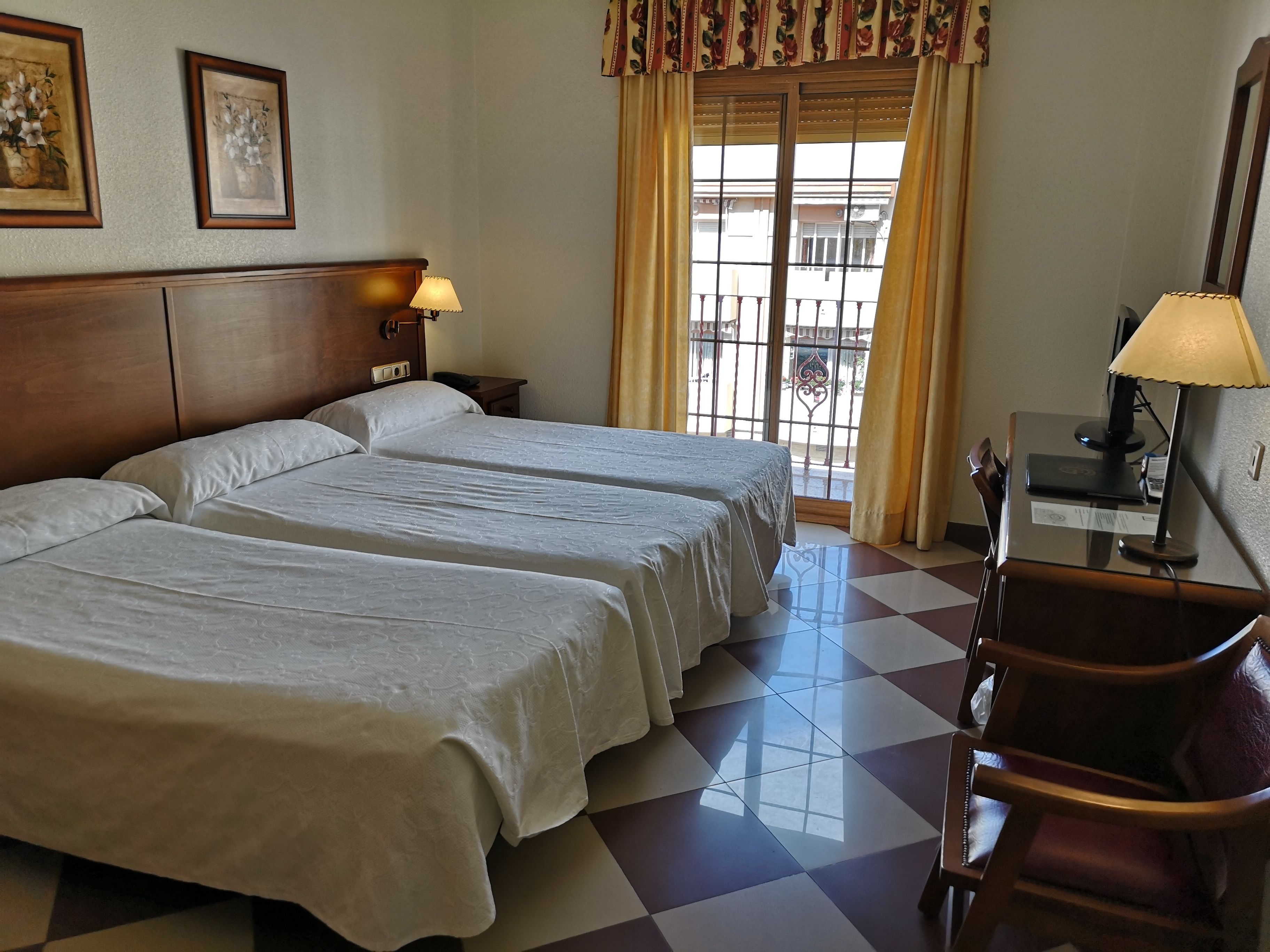 Habitación de hotel con dos camas, un escritorio, una silla, una lámpara y un ventanal con cortinas.