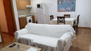 Una sala de estar con un sofá, una mesa con un control remoto y un folleto, un comedor con sillas y una mesa, una cocina con electrodomésticos y un cuadro en la pared.