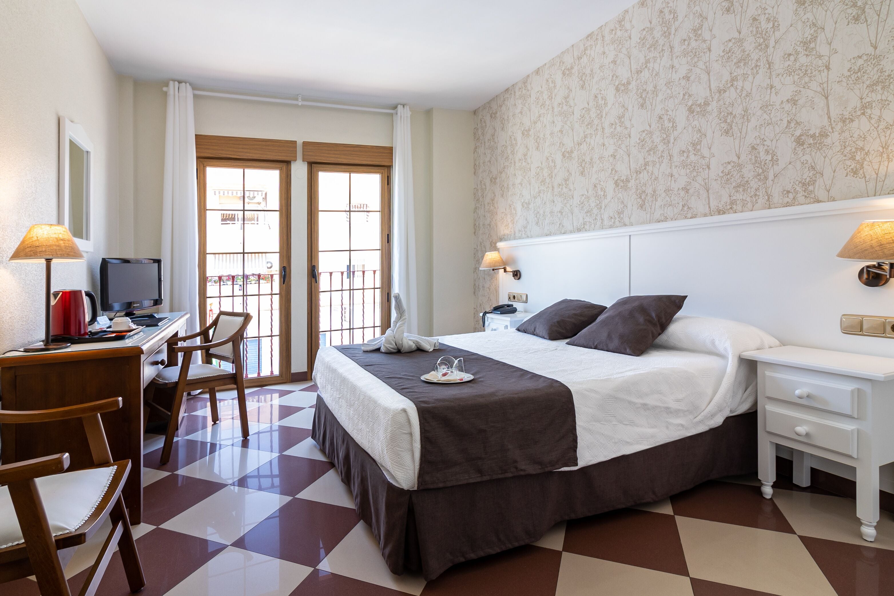 Habitación de hotel con piso a cuadros, una cama con colcha marrón, un escritorio con computadora y lámpara, y una ventana con cortinas.