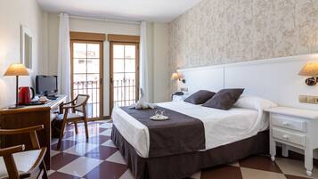 Habitación de hotel con piso a cuadros, una cama con colcha marrón, un escritorio con computadora y lámpara, y una ventana con cortinas.