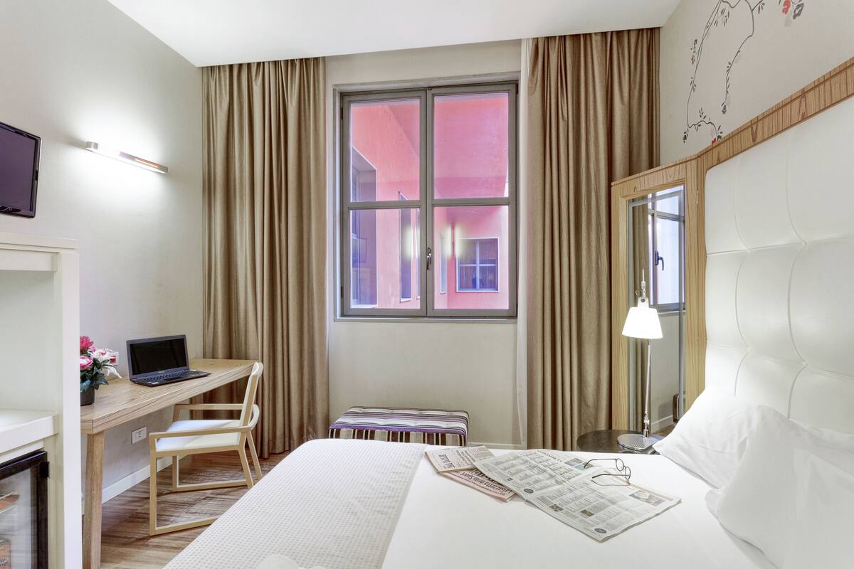 Une chambre d’hôtel comprenant un lit, un bureau avec un ordinateur portable, une chaise, une petite table, un miroir et une fenêtre avec des rideaux.