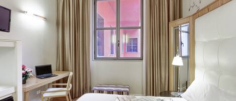 A hotel room with a bed, a desk with a laptop, a chair, a small table, a mirror, and a window with curtains.