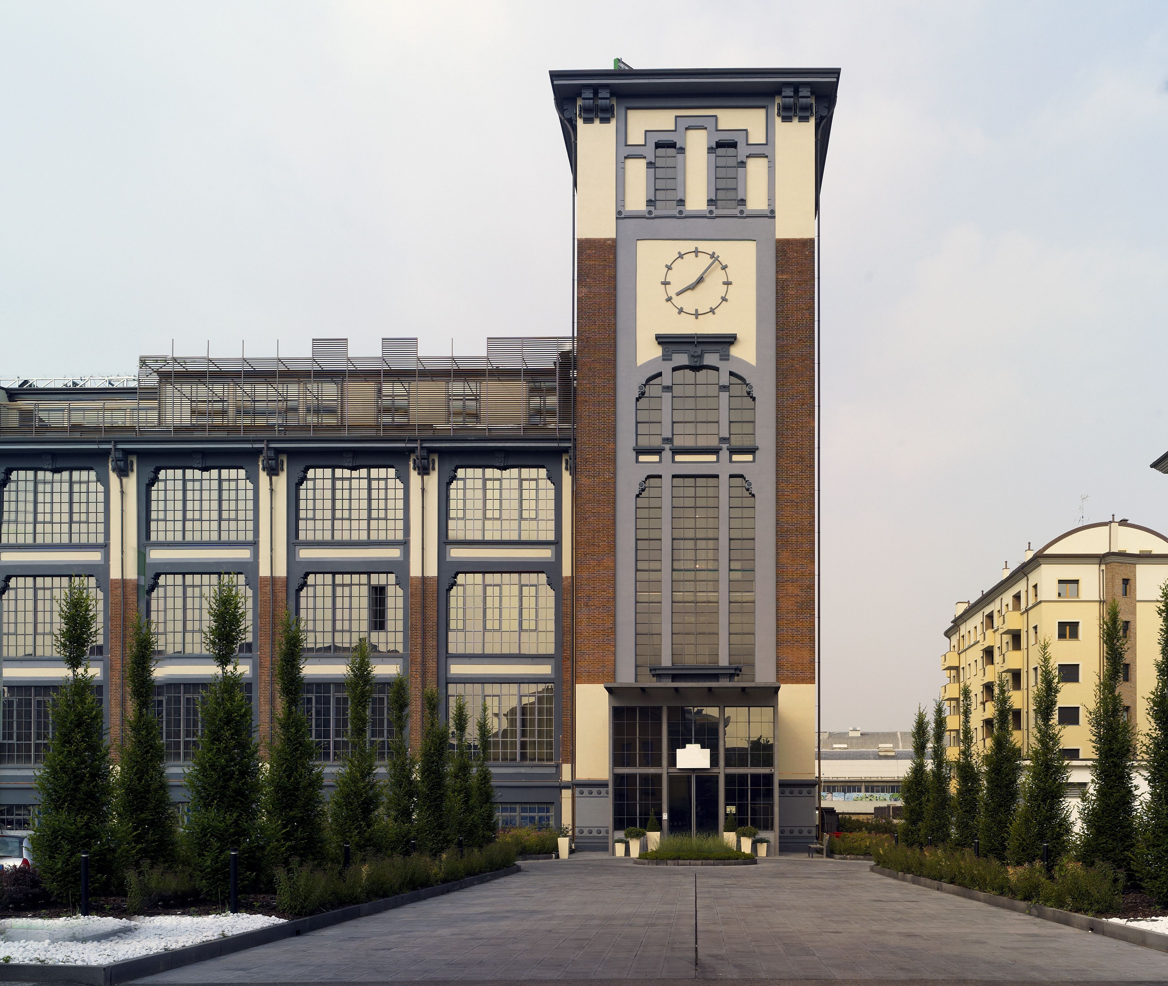 A modern building with a prominent clock tower, large windows, and a paved entrance area with landscaping.