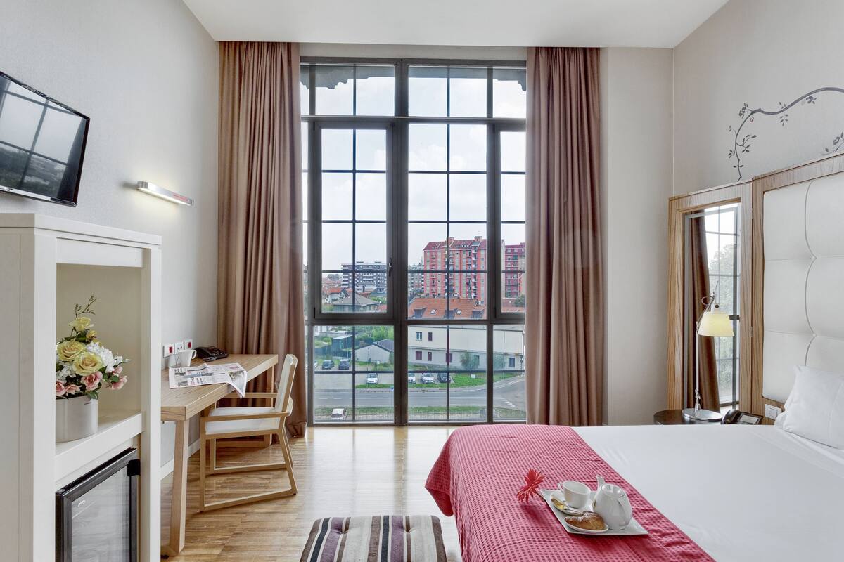 Une chambre d’hôtel avec une grande fenêtre, un lit, un bureau avec une chaise, une télévision et une petite table sur laquelle se trouve un plateau avec des articles pour le petit-déjeuner.