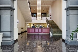 A hotel lobby with a reception desk, marble flooring, a staircase, and a large chandelier.