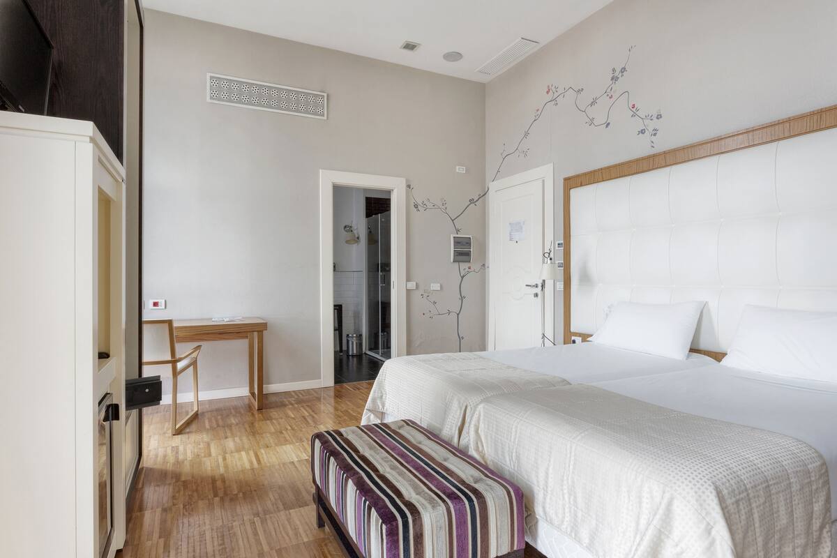 Une chambre à coucher comprenant un grand lit, une table de chevet, un banc, une armoire et une porte donnant sur une salle de bain.